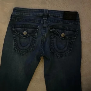 Mörkblå jeansbyxor från True Religion - Säljer ett par klassiska mörkblå jeans från True Religion med ikoniska bakfickor och snygga kontrastsömmar. Byxorna har normal passform och raka ben, samt knappgylf. Storlek 27 i uk storlekar som alltså är typ 32/34 kan funka för 36