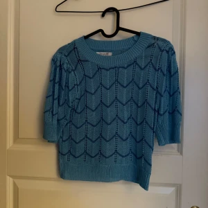 Blå stickad topp med mönster  - Säljer en ljusblå stickad topp med korta ärmar och ett öppet, hålmönstrat zigzag-mönster. Tröjan är croppad och har ribbade kanter vid hals, ärmslut och nederkant. Perfekt för vår och sommar!