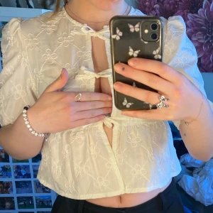 Vit blommig blus med rosetter - Supersöt vit blus med broderade blommor och tre små rosetter framtill. Blusen har korta puffärmar och är croppad, perfekt till både jeans och kjol. Ger en romantisk och trendig look.