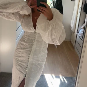 Vit långklänning från adoore  - Superfin vit långklänning med v-ringning, puffiga långärmar och dekorativa knappar framtill. Klänningen har en slits framtill och ett diskret blommigt mönster i tyget. Perfekt för sommarens speciella tillfällen. ALDRIG använd endast testad med lappar kvar!🤍🤍🤍🤍