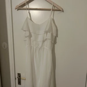 Vit klänning  - Säljer en söt vit klänning med smala axelband och volangdetaljer upptill. Klänningen har en luftig passform och är perfekt för varma dagar. Enkel och stilren design som passar till många tillfällen.