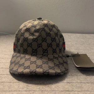  Gucci keps - Snygg beige keps från Gucci med klassiskt GG-monogram i mörkblått och röd-blå rand på sidan. Justerbar rem baktill i skinn och böjd skärm. Perfekt accessoar för en trendig look. Storlek ”M”