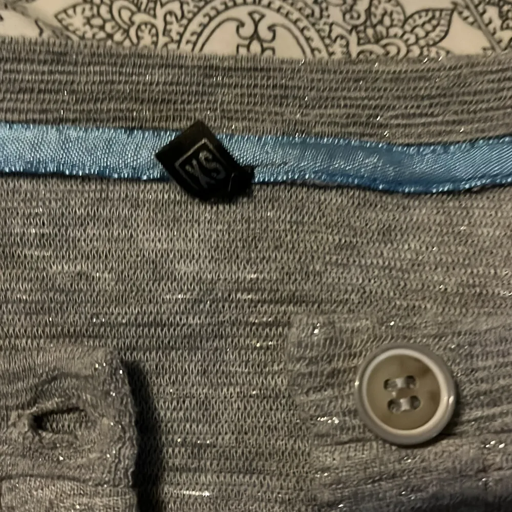 Säljer en grå croppad kofta med diskret glittrigt material och knappar framtill. Koftan har långa ärmar och en enkel, stilren design. Perfekt att slänga på sig över en topp för en extra touch av glitter. Koftan har relativt korta ärmar.. Neuletakit & Villapaidat.