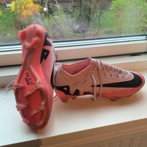 Nike Mercurial fotbollsskor i vitt och rosa Replicas - Säljer ett par Nike Mercurial fotbollsskor i vitt och rosa med svart swoosh. Skorna har snörning, platt sula med dobbar och är tillverkade i syntetmaterial. Perfekta för dig som vill ha lätta och snabba skor på planen. Pris kan diskuteras😁
