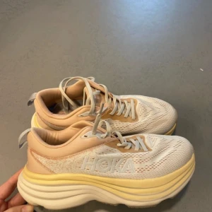 Hoka Bondi 8 sneakers i beige och vitt och ljusrosa - Säljer ett par Hoka Bondi 8 sneakers i beige och vitt med inslag av ljusgult. Skorna har en tjock, dämpad sula och ventilerande mesh-ovandel. Perfekta för dig som vill ha bekväma och snygga träningsskor med modern design.