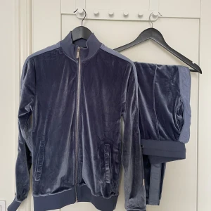 Mörkblått velourset med dragkedja - Säljer ett mörkblått veloursetfrån Maniére De Voir med dragkedja framtill och ribbade muddar. Tröjan har en sportig look med ståkrage och är tillverkad i mjuk polyesterblandning. Perfekt för en avslappnad och trendig stil. Mått byxor: 68 cm midja (stretch), 95cm längd utsida, 73cm längd insida Överdel: ärmlängd armhåla ner 52cm, bröstvidd 40 cm, längd 64cm (axel och ner)