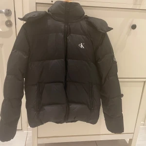 Svart dunjacka från Calvin Klein Jeans - Säljer en svart dunjacka från Calvin Klein Jeans med huva och dragkedja. Jackan har en klassisk pufferstil och logga på bröstet. Perfekt för kyliga dagar och enkel att matcha med olika outfits.