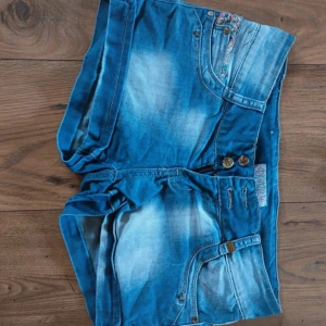 Blå jeansshorts med knappar - Snygga blå jeansshorts med slitna detaljer och dubbla knappar framtill. Klassisk femficksmodell och lågmidjad passform. Perfekta för sommaren och enkla att matcha med olika toppar.