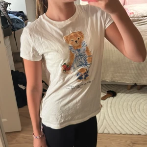 Vit t-shirt med björn från Polo Ralph Lauren - Vit t-shirt från Polo Ralph Lauren med en söt björn i blå klänning och sneakers tryckt på framsidan. Klassisk rund hals och korta ärmar. Materialet är mjuk bomull som känns skönt mot huden. Perfekt för dig som gillar gulliga och stilrena plagg med en twist.
