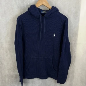 Marinblå stickad hoodie från Polo Ralph Lauren - Säljer en marinblå stickad hoodie från Polo Ralph Lauren med klassisk huva och snörning. Tröjan har en broderad vit logga på bröstet och en stor magficka. Perfekt för dig som gillar stilren och chill look.