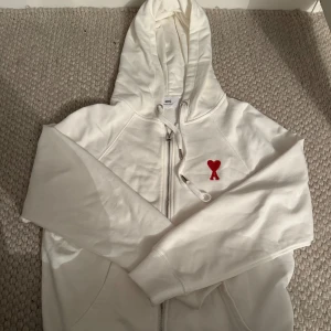 Vit hoodie med rött hjärta från Ami - Vit hoodie från Ami med dragkedja och huva. Tröjan har ett broderat rött hjärta med bokstaven A på bröstet, snörning vid huvan och långa ärmar. Perfekt för en chill och stilren look.