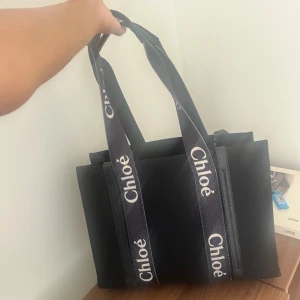 Svart shoppingväska från Chloé - Snygg svart shoppingväska från Chloé med breda axelband i textil där loggan är tryckt i vitt. Väskan har en boxig form och är tillverkad i ett kraftigt tygmaterial. Perfekt för dig som vill ha en trendig och rymlig väska med lyxig känsla.