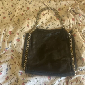 Svart axelväska med kedjedetaljer Tiamo - Snygg svart axelväska från Tiamo i skinnimitation med grova silvriga kedjor längs kanterna och som handtag. Väskan har dragkedja både på utsidan och insidan, perfekt för att hålla koll på dina grejer. Stilren och edgy design som lyfter vilken outfit som helst. Säljer för att den inte kommer till användning. Liten defekt därför så lågt pris.