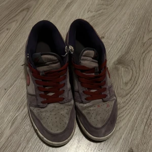 Nike Dunk sneakers i grått och rött - Nike Dunk sneakers med grå mocka och röda detaljer. Skorna har röda snören, röd Swoosh och mörkblå insida. Klassisk låg modell med perforerad tå och platt sula. Perfekta för dig som gillar retrovibbar och streetstyle.