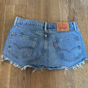 Levi's blå jeansshorts med fransar - Snygga blå jeansshorts från Levi's med klassisk femficksdesign och rå, fransig kant nedtill. De har hög midja och en liten broderad blomdetalj på ena bakfickan. 