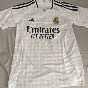 Real Madrid Mbappé 9 Adidas tröja M - Snygg vit Real Madrid fotbollströja från Adidas med Mbappé och nummer 9 på ryggen. Tröjan har svarta detaljer, klubbmärke på bröstet och sponsortryck framtill. Tillverkad i lätt och ventilerande Climalite-material, perfekt för match eller träning.