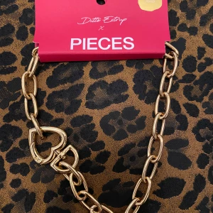 Guldfärgat halsband Pieces x Ditte Estrup - Snyggt halsband från Pieces x Ditte Estrup, 18K guldpläterat med chunky länkar och hjärtformad låsdetalj. Perfekt statement piece som lyfter din outfit med sin glansiga guldfärg och trendiga design.