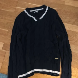 Marinblå stickad tröja med v-ringning - Snygg marinblå stickad tröja från Marine med v-ringad hals och vita ränder vid halsringning och nederkant. Tröjan har kabelstickat mönster och långa ärmar, perfekt för en preppy look. Passar dig som gillar klassisk stil med sportig touch.