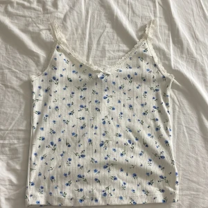 Vit blommig linnetopp från H&M Divided - Supersöt vit linnetopp med små blå blommor och tunna vertikala ränder. Toppen har spetskant vid axelbanden och en liten rosett framtill. Perfekt för varma dagar och ger en romantisk vibe till din outfit.