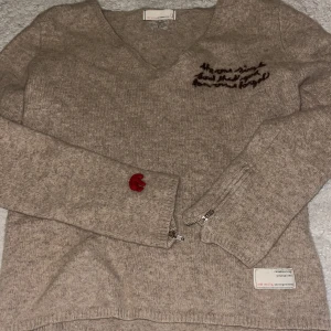 Beige stickad Odd Molly tröja v-ringad - Beige stickad tröja från Odd Molly med v-ringning, broderad text på bröstet och en röd broderad rosett på ärmen. Tröjan har långa ärmar, dragkedja vid ärmslut. Inga defekter 💞