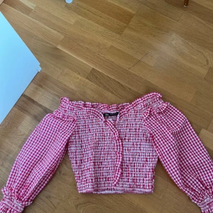 Rutig offshoulder blus från Zara - Supersöt röd och vit rutig blus från Zara med offshoulder och långa puffärmar. Smockad över hela kroppen och ärmarna, volangdetaljer vid halsringningen och en croppad passform. Perfekt för dig som gillar romantisk och trendig stil.