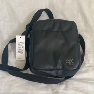 Svart axelväska från Calvin Klein - Minimalistisk svart axelväska från Calvin Klein Jeans med stilren CK-logga i metall på framsidan. Väskan har en rektangulär form, justerbar axelrem och är tillverkad i slätt skinnmaterial. Perfekt för att bära dina essentials och ge din outfit en clean vibe.