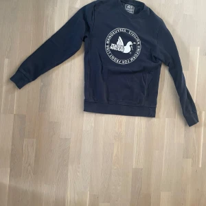 Blå sweatshirt från Peaceful Hooligan - Blå sweatshirt från Peaceful Hooligan med stort vitt tryck på bröstet och rund hals. Tröjan har långa ärmar och ribbade muddar vid ärmslut och nederkant. Perfekt för dig som gillar streetwear och vill ha en clean look med tydlig logga.