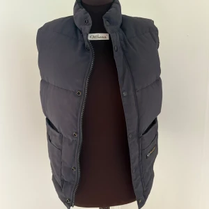 Svart dunväst från Canada Goose - Säljer en svart dunväst från Canada Goose med hög krage och stora fickor framtill. Västen har dragkedja och tryckknappar, samt en klassisk logotyp på fickan. Perfekt för lager-på-lager och chill höst- eller vinterstil. (Storleken är 2xs men passar mig som är runt s) 