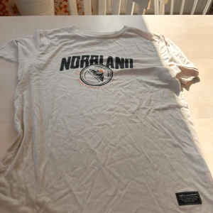 Vit t-shirt Norrland SQRTN Company - Vit t-shirt från SQRTN Company med tryck 'NORRLAND' och grafiskt motiv framtill. Klassisk rund hals och korta ärmar. Skön bomullskvalitet, perfekt för dig som gillar streetwear med norrländsk vibe.