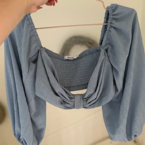 Blå blus med rosett från NA-KD - Supersöt ljusblå blus från NA-KD med stora puffärmar och en snygg rosettdetalj framtill. Toppen har smockad rygg och är tillverkad i ett mjukt, strukturerat tyg. Perfekt för dig som vill sticka ut med en trendig och feminin look.