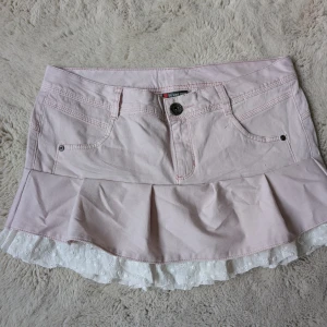 Egen sydd Ljusrosa volangkjol  - Supersöt ljusrosa kortkjol med volangkant och vit spetsdetalj längst ner. Kjolen har klassiska jeansfickor fram och bak, knapp och dragkedja i midjan. Perfekt för dig som gillar romantisk och feminin stil. Kom privat för mått och bild på