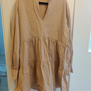 Beige volangklänning med v-ringning - Säljer en beige klänning med v-ringad hals och volangdetaljer. Klänningen har lång ärm och är tillverkad i ett lätt bomullsmaterial. Den har en loose passform och är perfekt för dig som gillar en avslappnad stil.