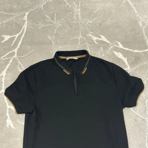 Svart piké LCW Vision med dragkedja - Svart pikétröja från LCW Vision i storlek M. T-shirten har korta ärmar och en krage med bruna detaljer samt en liten dragkedja framtill. Materialet känns som bomull och passformen är normal. Perfekt för dig som gillar stilrena och klassiska plagg med en twist.