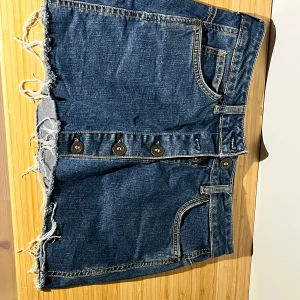 Blå jeanskjol med knappar - Säljer en blå jeanskjol från Gina tricot i storlek 36. Kjolen är kort med fransig kant nedtill och har flera fickor samt knappar framtill. 