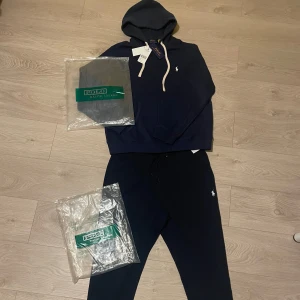 Marinblå set från Polo Ralph Lauren - Snygg marinblå hoodie och byxor från Polo Ralph Lauren med vit broderad logga på plaggen! Tröjan har huva med snörning, dragkedja framtill och långa ärmar. Byxorna har en sportig feel med skönt material. Tillverkad i mjuk bomullsmix som är perfekt för chill dagar. Klassisk och stilren design .
