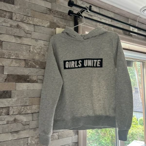 Grå hoodie från Gina Tricot med tryck - Snygg grå hoodie från Gina Tricot i storlek M med texten 'GIRLS UNITE' i svart och vitt på bröstet. Tröjan har huva och långa ärmar, tillverkad i mjuk bomullsmix som är perfekt för chill dagar. Klassisk passform och ribbade muddar vid ärmslut och nederkant.