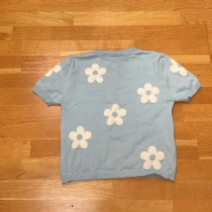 Blå stickad tröja med blommor - Supersöt ljusblå stickad tröja med korta ärmar och vita blommor som är spridda över hela tröjan. Ribbad kant vid hals, ärmslut och nederkant. Perfekt för dig som gillar retrovibes och vill ha något som sticker ut lite extra i garderoben.