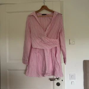 Rosa och vit randig skjortklänning - Säljer en rosa och vit randig skjortklänning med omlott-detalj och knappar framtill. Klänningen har lång ärm, klassisk krage och ett knytband i midjan som ger en snygg siluett. Perfekt för dig som gillar en fräsch och feminin stil.