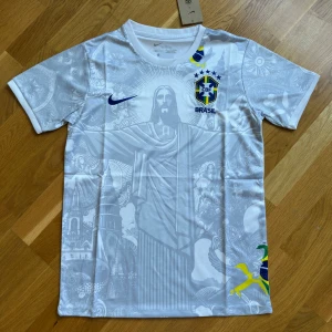 Vit Brasil Nike fotbollströja - Snygg vit Brasilien fotbollströja från Nike med korta ärmar och detaljerat grafiskt tryck på framsidan, inklusive Kristusstatyn och brasilianska motiv. CBF-emblem och Nike-logga på bröstet, samt brasilianska färger på nederkanten. Perfekt för fans av landslaget.