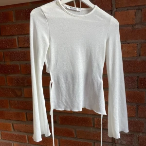 Vit långärmad topp från NA-KD - Säljer en vit långärmad topp från NA-KD i storlek XS. Toppen har en rund halsringning, croppad passform och snygga knytband i sidan som ger en cool detalj. Materialet är mjukt och lätt, perfekt för vår och sommar. Enkel att styla med jeans eller kjol.