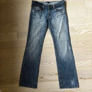 Blå Lågmidjade bootcut jeans med slitningar - Snygga blå lågmidjade jeans med bootcut-modell och slitna detaljer framtill och baktill. Jeansen har klassiska fem fickor, bälteshällor och dragkedja med knapp. Materialet är robust denim som ger en cool, avslappnad vibe. Storleken är S/M