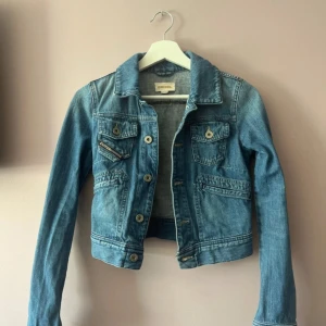 Blå jeansjacka från Diesel - Snygg blå jeansjacka från Diesel med klassisk krage, knappar framtill och flera fickor med både knapp och dragkedja. Jackan har en croppad passform och är tillverkad i jeansmaterial. Perfekt för dig som vill ha en cool och tidlös look.