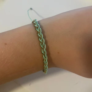 Flätat turkost fotlänk/armband med gulddetaljer - Snyggt armband i turkost med flätad design och små guldfärgade pärlor längs hela bandet. Justerbar knytning Perfekt accessoar för en färgglad och personlig stil. Egentligen en gotlänning men kan användas som armband också.