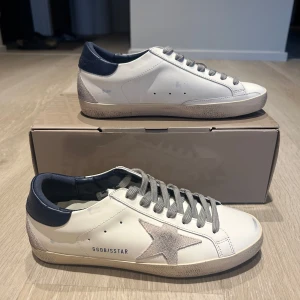 Golden Goose vita sneakers med grå stjärna - Golden Goose sneakers i vitt skinn med grå mockadetaljer och ikonisk stjärna på sidan. Grå snörning, mörkblå häl och beige sula med vintage-look. Perfekta för dig som gillar en clean men ändå edgy stil. Kommer med originalpåse och certifikat.