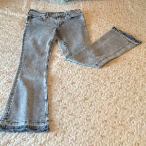 Ljusblå bootcut jeans med rå kant - Säljer ett par ljusblå bootcut jeans med snyggt slitna och råa kanter nertill. Jeansen har klassisk femficksdesign, dragkedja och knapp framtill. Perfekt tvättad denim med coola detaljer längs bensluten. Passar dig som gillar retrovibbar och avslappnad stil. Längd från midjan ner: 85 cm. Midjan: 31,5 cm