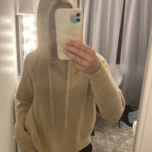 Beige hoodie i stickat material - Mysig beige hoodie med huva och snörning framtill. Tröjan är stickad och har en loose passform med långa ärmar. 