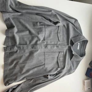 Grå overshirt från Zara XL - Stilren grå overshirt från Zara i storlek XL. Skjortan har två stora bröstfickor med lock, klassisk krage och knäppning framtill. Perfekt lager-på-lager-plagg med lång ärm och enkel, clean look.