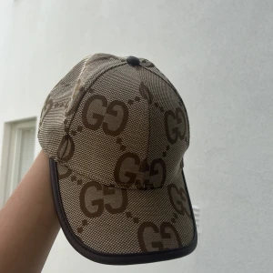  beige keps med GG-mönster - Snygg keps i beige och brunt med ikoniskt GG-mönster över hela kepsen. Kepsen har böjd skärm, bruna detaljer och justerbar rem bak i läder. Perfekt för dig som vill sticka ut med en lyxig accessoar.