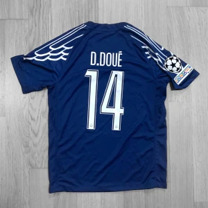 PSG blå matchtröja D. Doué #14 - Snygg blå PSG fotbollströja med D. Doué och nummer 14 på ryggen. Tröjan har vita detaljer, vingmönster på axlarna, Qatar Airways tryck på bröstet och Champions League-patch på ärmen. Tillverkad i lätt och ventilerande polyester, perfekt för match eller träning.
