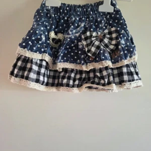 Blå och vit prickig och rutig kjol - Supersöt kortkjol med blått tyg med vita prickar upptill och svartvit rutigt tyg nedtill. Kjolen har volanger, spetsdetaljer längs kanterna, en stor rutig rosett och ett hjärta i spets. Perfekt för dig som gillar unika detaljer och mixade mönster. (Det står storlek XXL på lappen, men det är fel. Den är inköpt på barnavdelningen och använts till flicka i storlek 92/98.)
